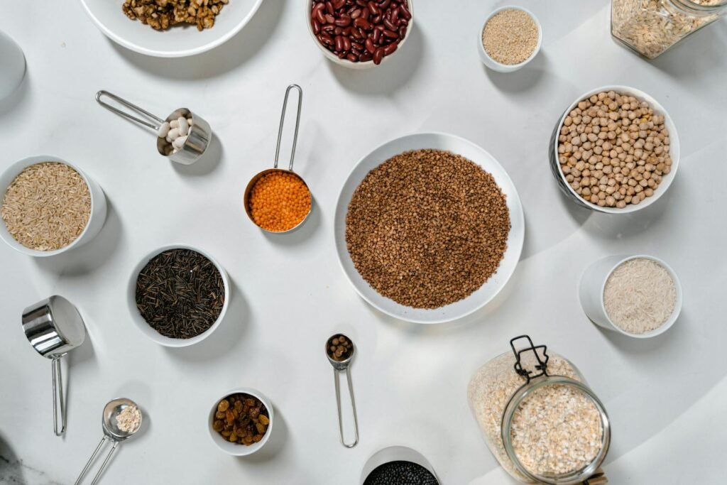 Top view of various grains, legumes, and spices in bowls and jars on a white table.